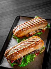 Two long submarine sandwiches with meat, cheese, bacon, tomatoes, lettuce, cucumbers and onions on black wooden table background