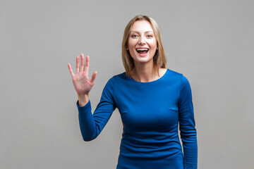 Hi! Portrait of sweet happy woman in elegant tight blue dress standing with raised hand saying hello to camera, greeting guests with friendly smile. indoor studio shot isolated on gray background