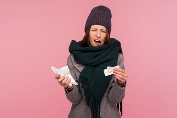 Upset depressed sick woman in hat and warm scarf holding napkin and pills, frowning with displeased miserable expression. Indoor studio shot isolated on pink background