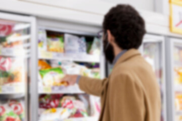 Young brunette man in a medical mask in the supermarket in the department with frozen foods. Coronavirus pandemic. Blurred.