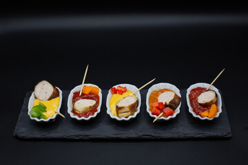 small plates with a piece of sausage in different dips with pepper pieces and wood skewer