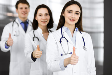 Group of young cheerful doctors is standing as a team with thumbs up in a hospital office and is...