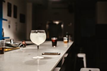 Una copa de alcohol con hielos junto a un teléfono móvil en la barra de un bar sin gente