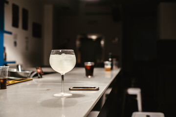 Una copa de alcohol con hielos junto a un teléfono móvil en la barra de un bar sin gente