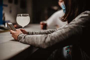 Una mujer joven sentada en la barra de un bar sosteniendo su teléfono móvil. En el mostrador hay un vaso de alcohol blanco con limón. La mujer lleva una mascarilla quirúrgica azul que cubre su boca