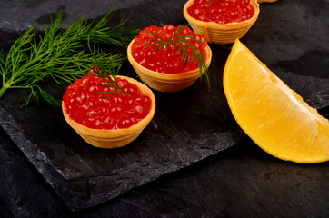Tartlets with red salmon caviar on black stone plate, decorated with dill and a slice of lemon, close - up. Seafood appetizer