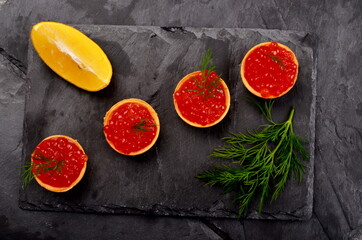 Tartlets with red salmon caviar on black stone plate, decorated with dill and a slice of lemon, close - up. Seafood appetizer