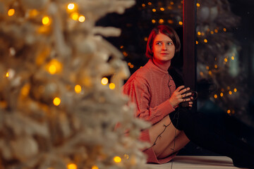 Beautiful young woman sits on window with Christmas decorations