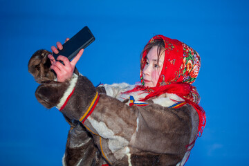 A young girl, in the national winter clothes of the northern inhabitants of the tundra, takes a selfie on a smartphone