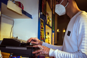 Un camarero joven tecleando en una caja registradora de un bar de noche. El hombre lleva puestos guantes y mascarilla como protección contra el virus