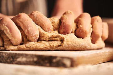 Female baker hands making dough for bread with an apron. Roller and chopping board visible in the background. Natural homemade ingredients. Dark background, brown color grading.