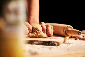 Female baker hands making dough for bread with an apron. Roller and chopping board visible in the background. Natural homemade ingredients. Dark background, brown color grading.