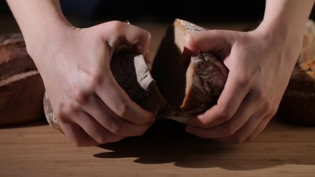 A fresh rustic bread loaf being ripped open by hand on wooden background. Tasty and healthy baked whole-wheat bread. 4K.