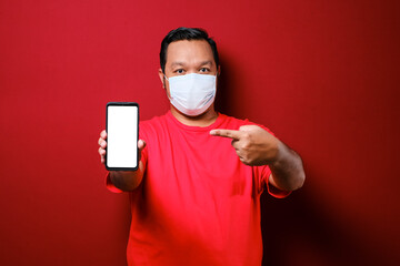 Excited young asian guy in medical mask showing blank white smart phone screen