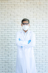 Portrait of Asian elder doctor or researcher wear lab coat, clear eyeglasses and face mask standing and arm crossed with white brick background.