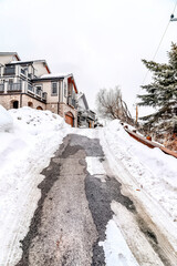 Road with guard rail going up a snowy slope leading to houses under white sky