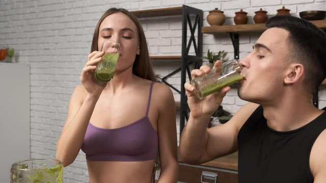 Making green juice at home: Young sporty couple in sportswear drinking smoothie from a glass. Close-up shot. Healthy breakfast after a workout in the kitchen 4k