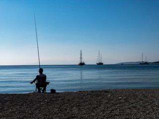 Fototapeta na wymiar A lonely fisherman silhouette sits in a chair at the beach and tries to catch some fishes. A cat lies beside him and waiting for food. Some sailing boats are on anchor. Blue sky and sea background