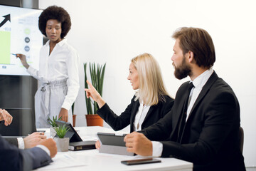 Male and female Caucasian business workers, listening to report of their African female colleague, standing in front of the big plasma digital wall screen with infographics charts. Focus on blond lady