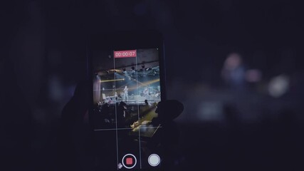 A close-up shot of a man's hand is taking a vertical video for a music band performing on stage and the neon light at the night club. - Powered by Adobe