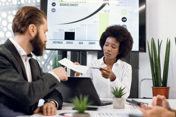 Young attractive African businesslady, showing some paper with infographic charts to her bearded high-skilled Caucasian male colleague at business meeting in modern office