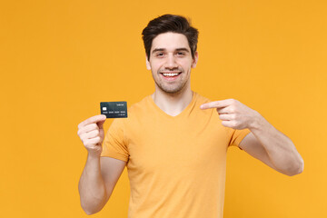 Young smiling unshaved successful caucasian man 20s in casual basic blank print design t-shirt hold in hand pointing index finger on credit bank card isolated on yellow background studio portrait.