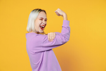 Young excited strong sporty woman bob haircut and open toothy mouth wearing casual basic purple shirt woman demonstrating muscles biceps on hand isolated on yellow color background studio portrait.