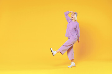Full length of young fun woman 20s bob haircut in casual basic purple suit beanie hat walk raised leg hold hand at forehead look far away distance isolated on yellow color background studio portrait.