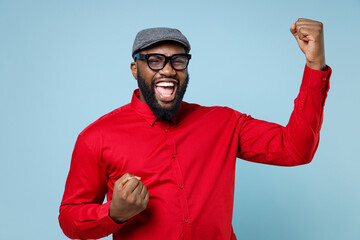 Naklejka premium Overjoyed happy young bearded african american man 20s wearing casual red shirt cap eyeglasses standing doing winner gesture clenching fists isolated on pastel blue color background studio portrait.
