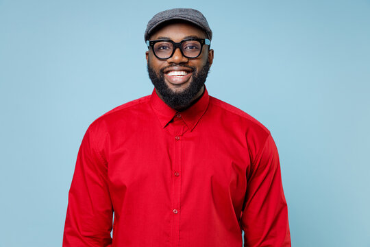 Cheerful smiling attractive young bearded african american man 20s wearing casual red shirt cap eyeglasses standing and looking camera isolated on pastel blue color wall background studio portrait.