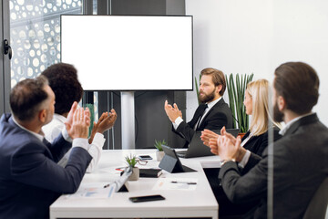 Happy group of five diverse multiethnic businesspeople clapping hands, while sitting at the table in modern office, in front of huge plasma TV screen with empty space for text