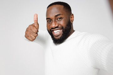Fototapeta premium Close up of smiling young african american man 20s in casual basic sweater doing selfie shot on mobile phone showing thumb up looking camera isolated on white color wall background studio portrait.