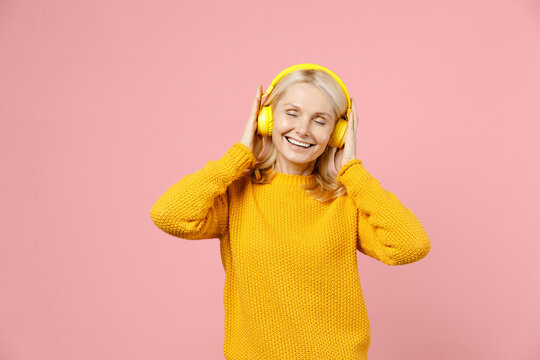 Cheerful elderly gray-haired blonde woman lady 40s 50s years old in yellow casual sweater listening music with headphones keeping eyes closed isolated on pastel pink color background studio portrait.