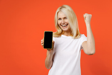 Happy elderly gray-haired blonde woman lady 40s 50s years old in white casual t-shirt hold mobile cell phone with blank empty screen doing winner gesture isolated on orange background studio portrait.