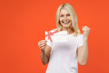 Happy elderly gray-haired blonde woman lady 40s 50s years old wearing white basic t-shirt hold gift certificate doing winner gesture looking camera isolated on orange color background studio portrait.