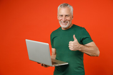 Smiling elderly gray-haired mustache bearded man wearing casual basic green t-shirt standing working on laptop pc computer showing thumb up isolated on bright orange color background studio portrait.