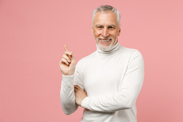 Smiling cheerful elderly gray-haired mustache bearded man wearing casual basic white turtleneck pointing index finger up on mock up copy space isolated on pastel pink color background studio portrait.