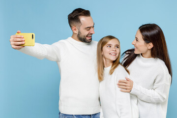 Pretty young parents mom dad with child kid daughter teen girl in white sweaters doing selfie shot on mobile phone hugging isolated on blue background studio. Family day parenthood childhood concept.