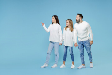 Fototapeta premium Full length of funny young parents mom dad with child kid daughter teen girl in sweaters holding hands pointing finger aside isolated on blue background studio portrait. Family day parenthood concept.