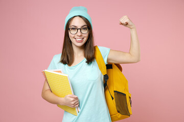 Strong smiling young woman student in basic blue t-shirt hat eyeglasses backpack hold notebooks showing biceps muscles isolated on pink background. Education in high school university college concept.