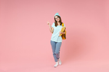 Full length portrait of funny young woman student in t-shirt hat glasses backpack hold notebooks showing victory sign isolated on pink background. Education in high school university college concept.