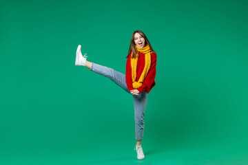 Full length side view of cheerful laughing young brunette woman 20s in casual knitted red sweater yellow scarf dancing standing rising leg isolated on bright green color background studio portrait.
