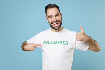 Funny young bearded man in volunteer t-shirt pointing index finger on himself showing thumb up isolated on blue background studio portrait. Voluntary free work assistance help charity grace concept.