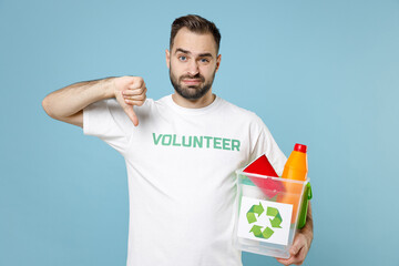 Displeased young man in white volunteer t-shirt hold basket with plastic trash showing thumb down...