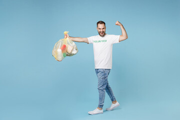 Full length strong young man in volunteer t-shirt hold bags with plastic paper trash showing biceps muscles isolated on blue background. Voluntary free assistance help trash sorting recycling concept.