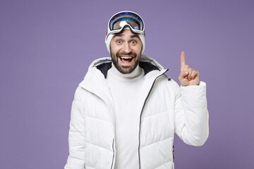 Excited skier man in white windbreaker jacket ski goggles mask hold index finger up with great new idea spend weekend winter in mountains isolated on purple background. People lifestyle hobby concept.