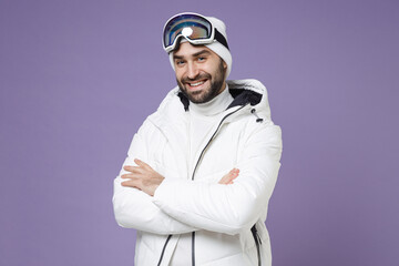 Smiling skier man in white windbreaker jacket ski goggles mask holding hands crossed spend extreme weekend winter season in mountains isolated on purple background. People lifestyle hobby concept.