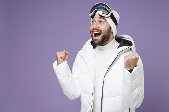 Excited skier man in warm white windbreaker jacket ski goggles mask doing winner gesture spend extreme weekend winter season in mountains isolated on purple background. People lifestyle hobby concept.