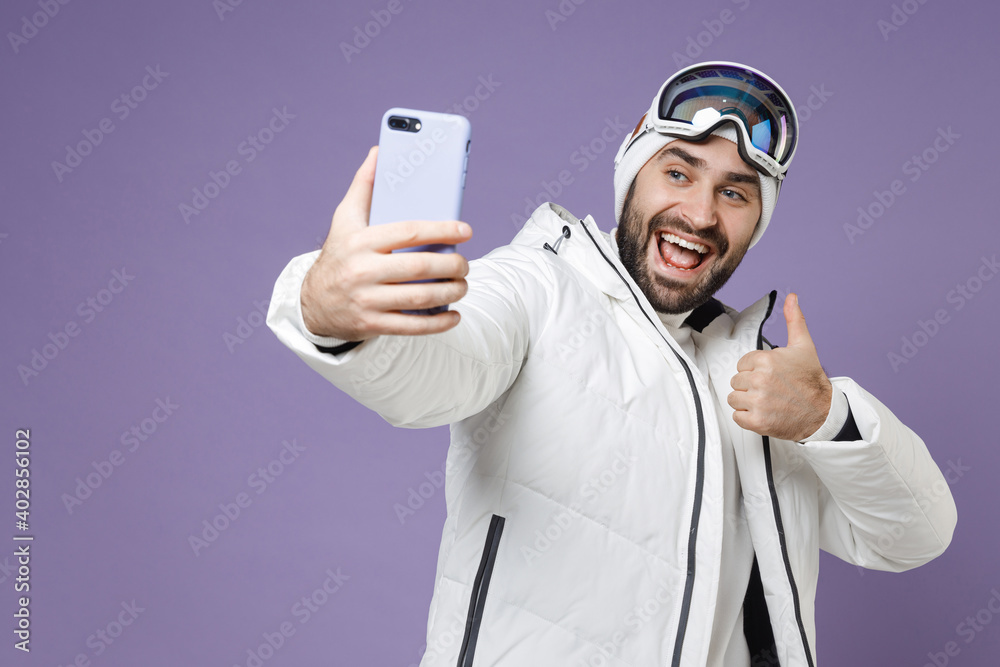 Wall mural cheerful skier man in white jacket ski goggles mask doing selfie shot on mobile phone showing thumb 