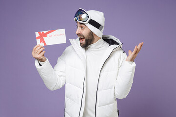 Surprised skier man in white windbreaker jacket ski goggles mask hold gift certificate spreading hands spend weekend winter in mountains isolated on purple background. People lifestyle hobby concept. © ViDi Studio